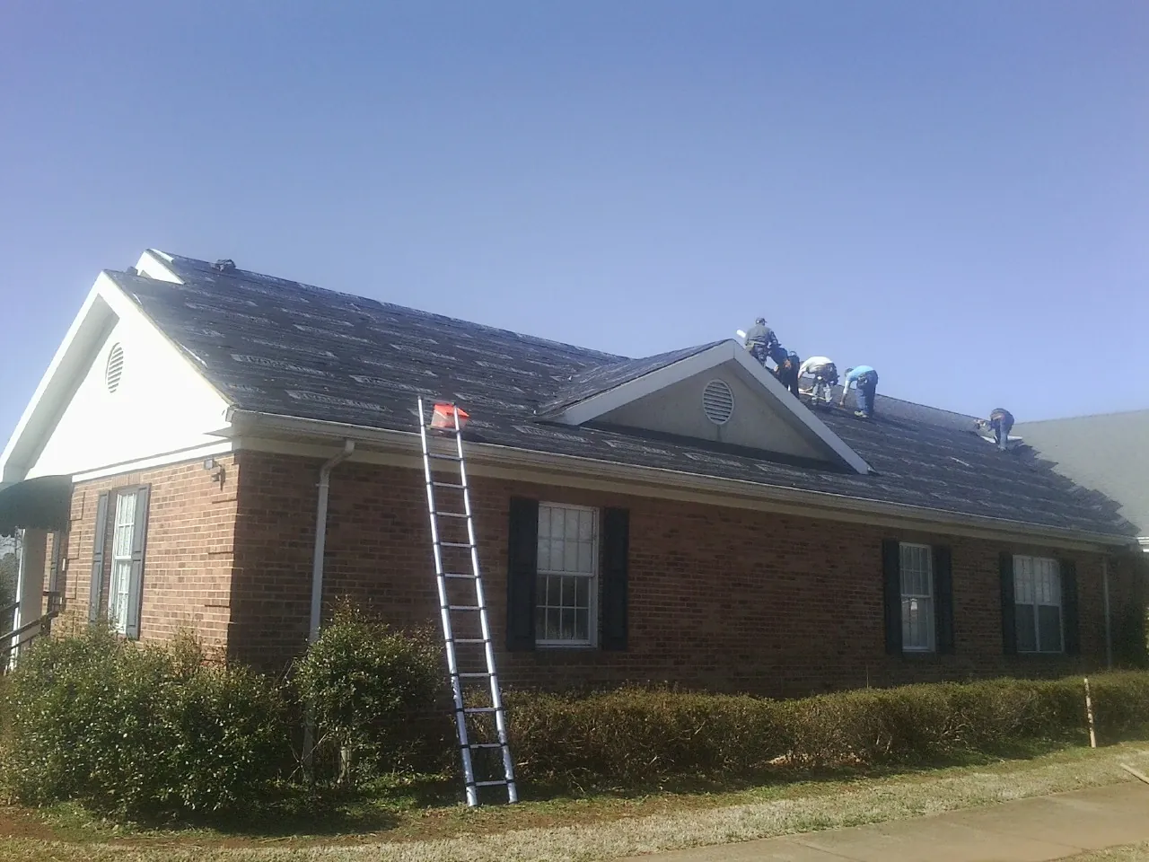 Crew tearing shingles off a brick ranch roof with a ladder set — Emergency Roof Repair work by our crew in Vader
