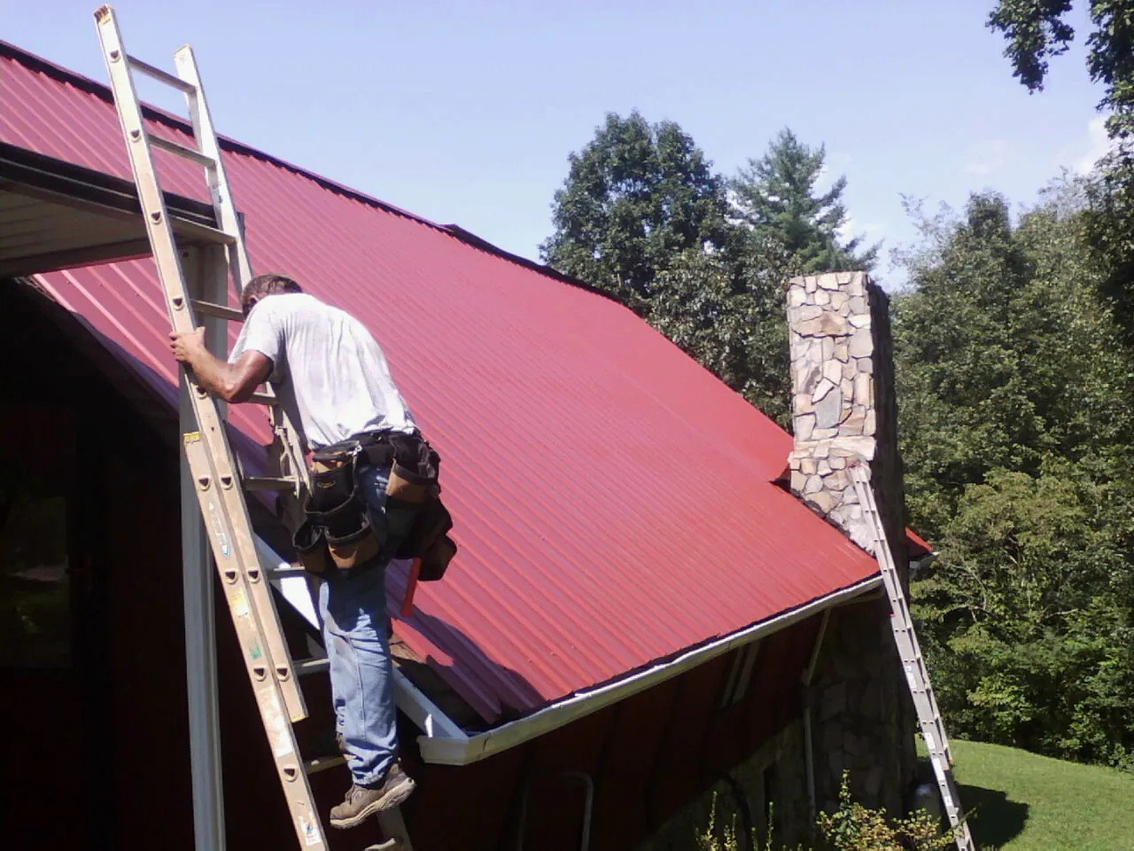 Roofer on a ladder beside a red metal roof and stone chimney — Soffit & Fascia Repair by Roofing Brothers in Vader